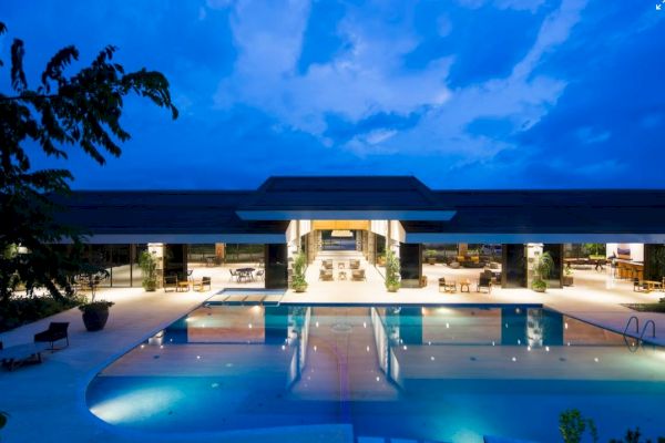 The image depicts a modern, illuminated building with a large outdoor swimming pool in front, surrounded by pool chairs and lush greenery.