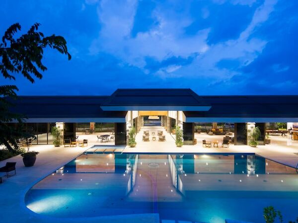 The image depicts a modern, illuminated building with a large outdoor swimming pool in front, surrounded by pool chairs and lush greenery.