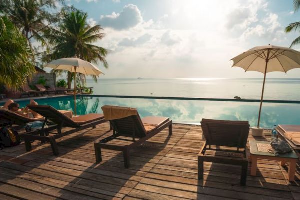 A serene poolside scene with lounge chairs, umbrellas, palm trees, and a calm sea under a partly cloudy sky at sunset.