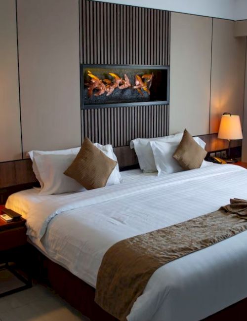 A cozy hotel room with a neatly made bed, two bedside tables with lamps, a small table, decorative pillows, and a curtain on the window.