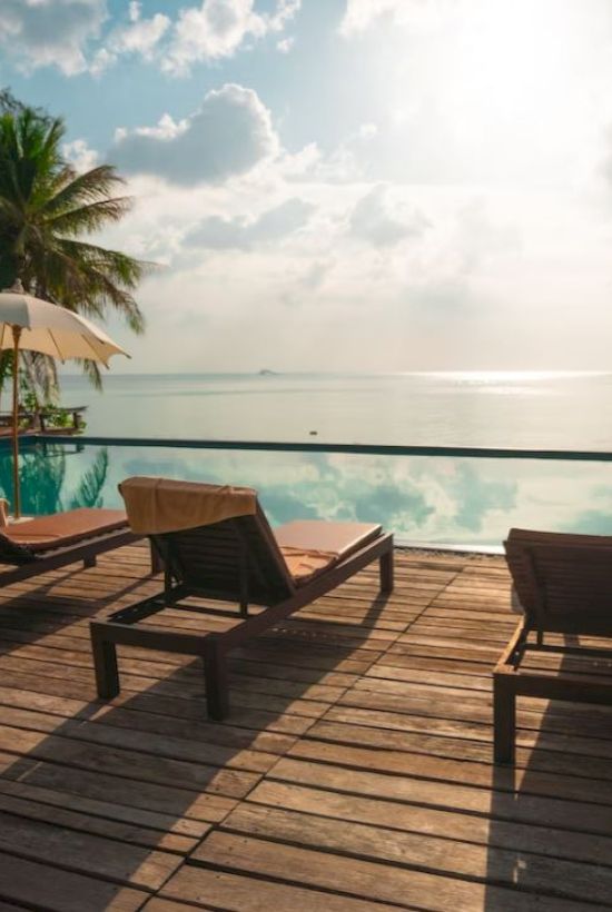 The image shows a serene poolside scene with lounge chairs, umbrellas, palm trees, and a reflective body of water under a sunny sky.