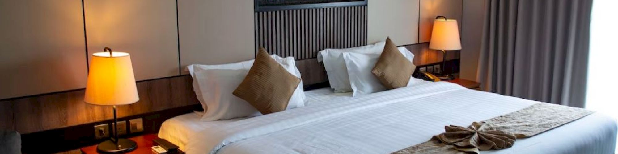 A well-decorated hotel room features a large bed with brown and white linens, two bedside lamps, and a wall-mounted decorative fireplace.