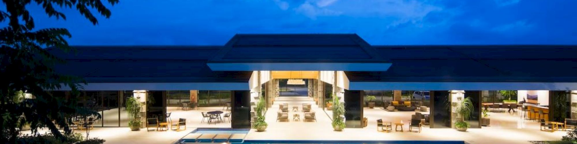 The image shows a luxurious villa with a large swimming pool, illuminated at night. Inside, there are various seating areas and tables.