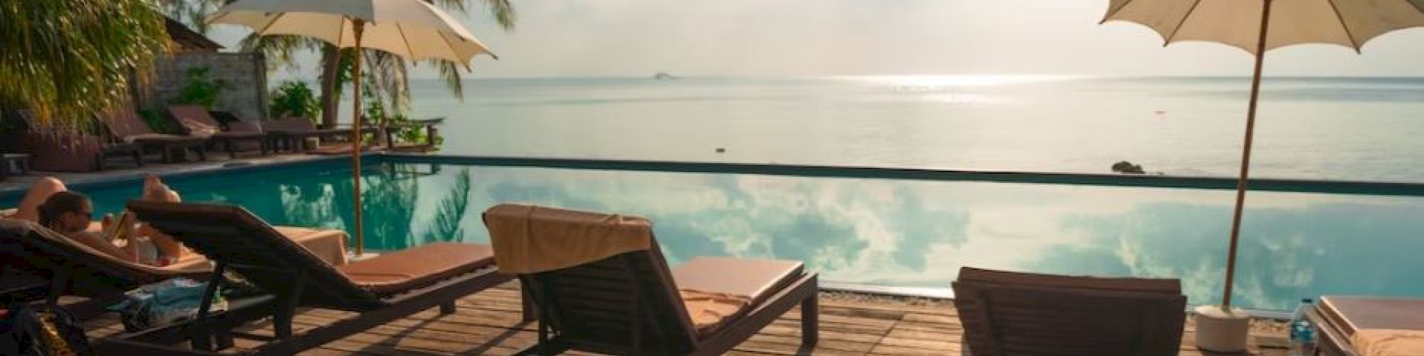 A serene poolside with lounge chairs, umbrellas, and palm trees, overlooking a calm ocean under a bright sky with scattered clouds.