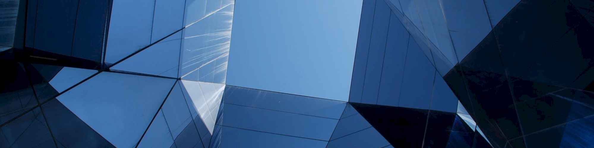 The image shows a view looking up at a geometric arrangement of reflective glass panels forming a tunnel-like structure with blue sky at the center of the frame.