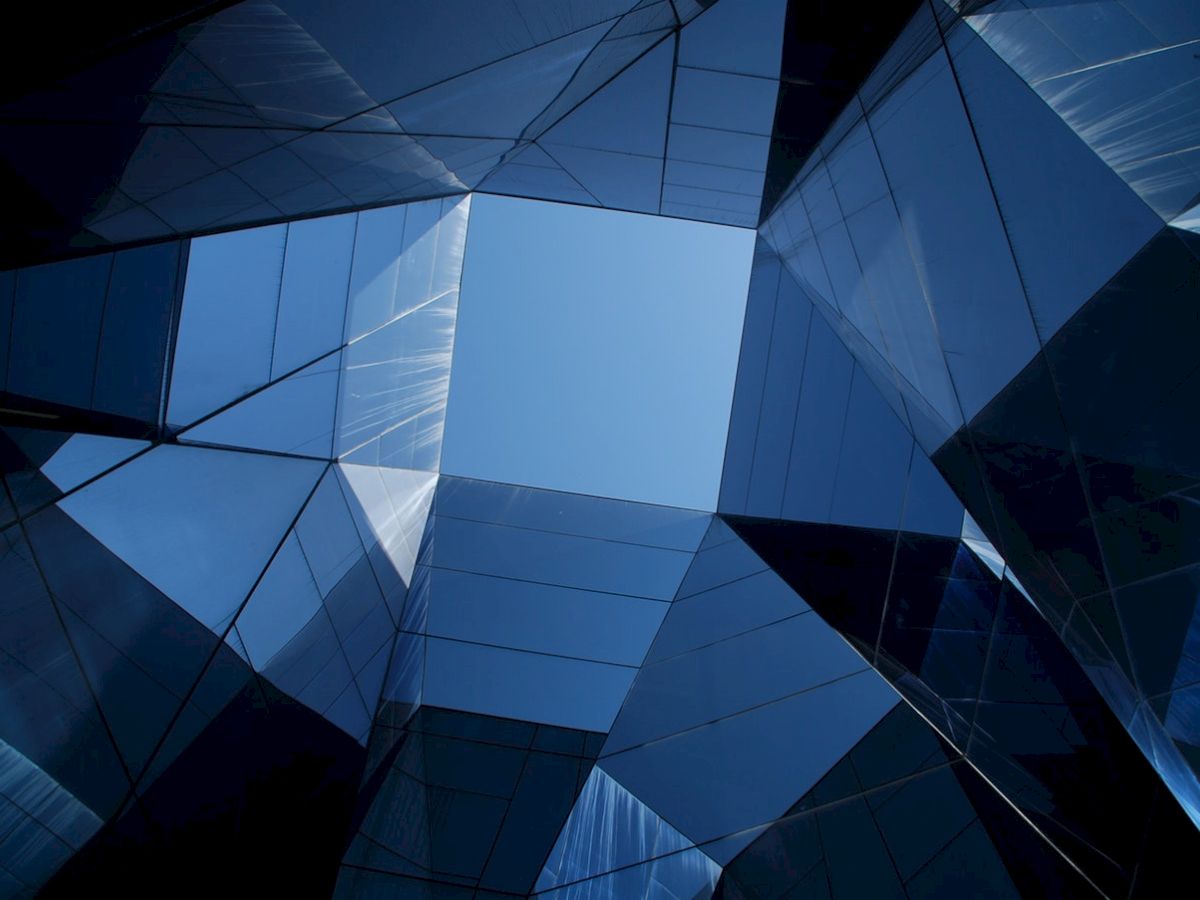 The image shows a geometric architectural structure with mirrored and glass panels reflecting the sky. The view is upward from within.