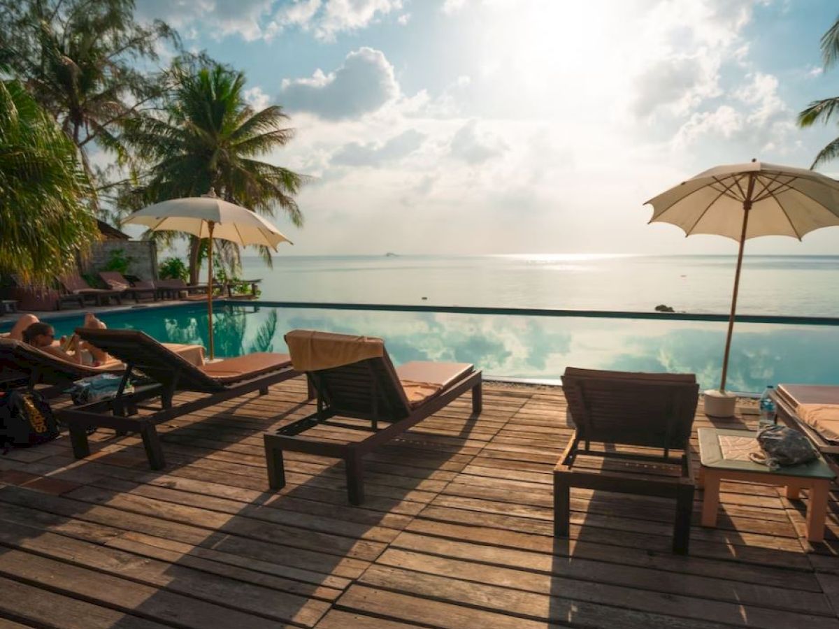 A serene beach scene with lounge chairs, umbrellas, and a beautiful view of the ocean from a wooden deck. The sky is partly cloudy and calm.