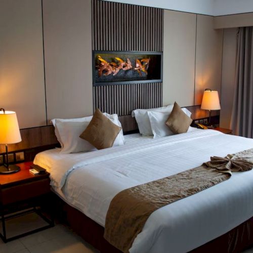 A well-lit hotel room with a large bed, two nightstands with lamps, and wall art above the headboard. Curtains cover a window on the right.