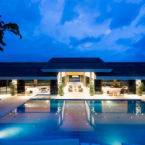 The image shows a modern building with large open spaces, a pool in the foreground, and illuminated interiors against a twilight sky.
