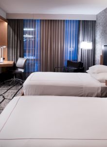 A modern hotel room with two beds, a wall-mounted TV, a desk, and a chair by the window. The room has stylish decor and ample lighting.