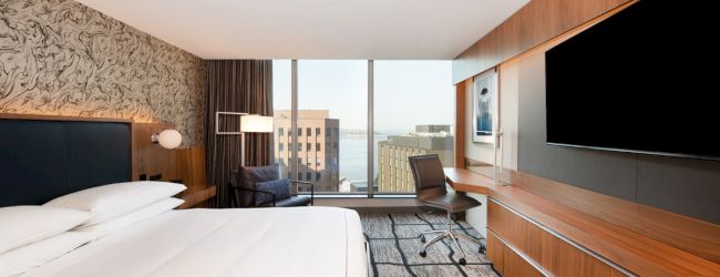 A modern hotel room features a large bed, wall-mounted TV, desk with chair, and large windows offering a city view in the background.