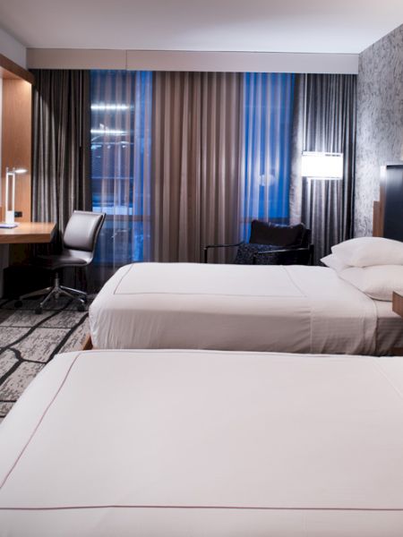 The image shows a modern hotel room with two double beds, a wall-mounted TV, a work desk, and contemporary decor, including patterned wallpaper and carpet.