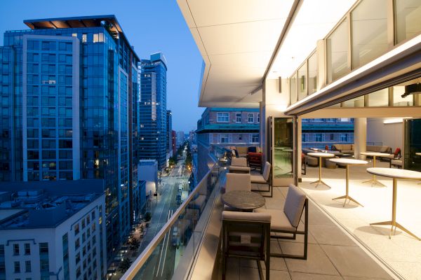 The image shows a modern cityscape with tall buildings and a stylish, partially enclosed terrace with seating, tables, and a view of the street below.