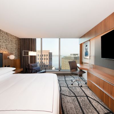 A modern hotel room with a large bed, wall-mounted TV, desk, chair, city view through large window, and stylish decor.