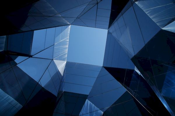 The image shows a view looking up through a geometric arrangement of mirrored or glass surfaces, reflecting a fragment of the blue sky.