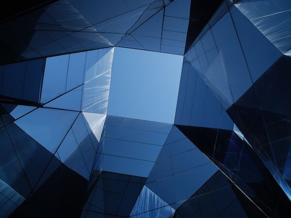 The image shows a view looking up through a geometric arrangement of mirrored or glass surfaces, reflecting a fragment of the blue sky.