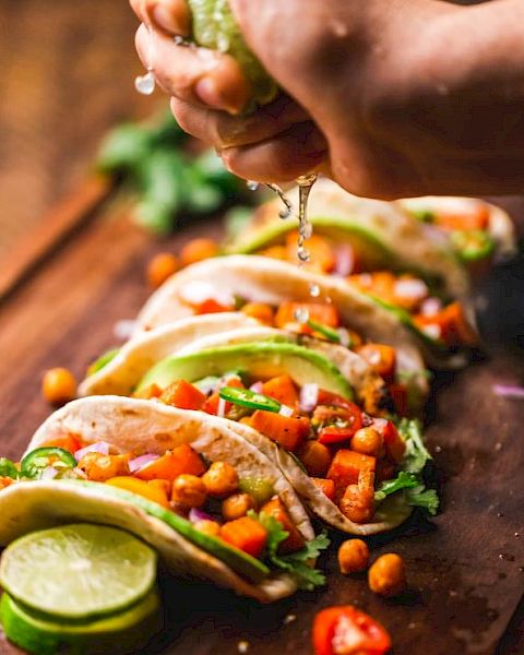 The image shows a close-up of a hand squeezing lime juice over a row of vegetable tacos garnished with chickpeas, cilantro, and avocado slices on a wooden board.