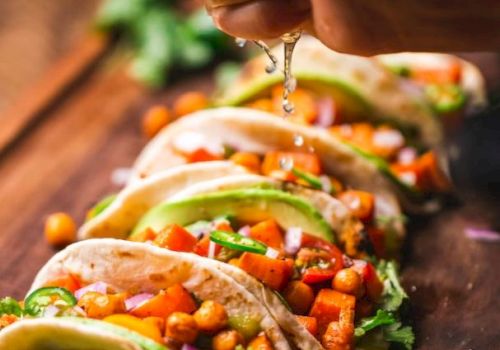 The image shows a close-up of a hand squeezing lime juice over a row of vegetable tacos garnished with chickpeas, cilantro, and avocado slices on a wooden board.