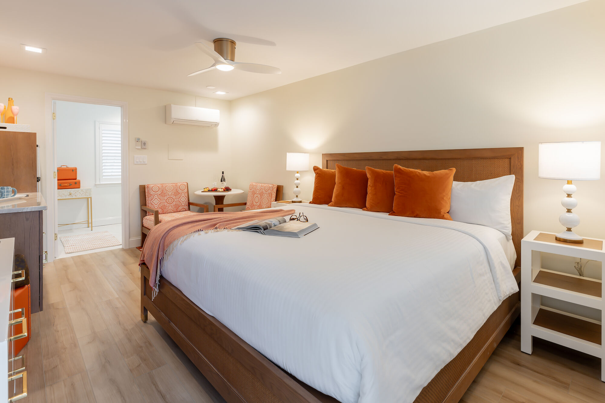 A neatly made bed with orange pillows, side tables with lamps, a ceiling fan, and a small dining area in a cozy bedroom with modern decor.