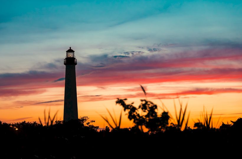 A lighthouse stands tall against a vibrant sunset sky with silhouettes of plants and trees in the foreground, creating a serene scene.