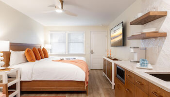 A cozy bedroom with a large bed, orange accents, TV, wooden shelves, white lamps, and a small kitchen counter with a sink and storage.