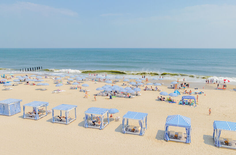 A beach scene with striped cabanas, umbrellas, and people enjoying the sand and ocean waves under a clear sky.