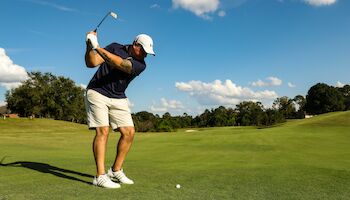 A person is playing golf on a lush green course, preparing to swing the golf club with a clear blue sky and trees in the background.