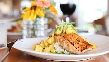 A plate of grilled salmon with garnishes of pineapple on a white plate, set on a table with flowers and a glass of red wine in the background.