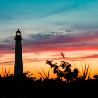 A lighthouse stands silhouetted against a vibrant sunset sky, with foliage visible in the foreground.