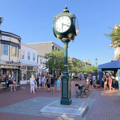 A town square with a green clock tower in the center, surrounded by people, benches, shops, and trees under a clear blue sky.