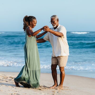 A couple is joyfully dancing on a sandy beach with the ocean waves in the background under a clear sky.