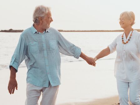 An older couple holding hands and walking along a beach, smiling at each other, with the sea in the background.