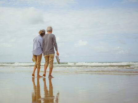 An elderly couple is walking barefoot on the beach, holding hands, and enjoying the view of the ocean under a partly cloudy sky.