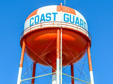 The image shows an orange and white water tower with "COAST GUARD" written on it against a clear blue sky.