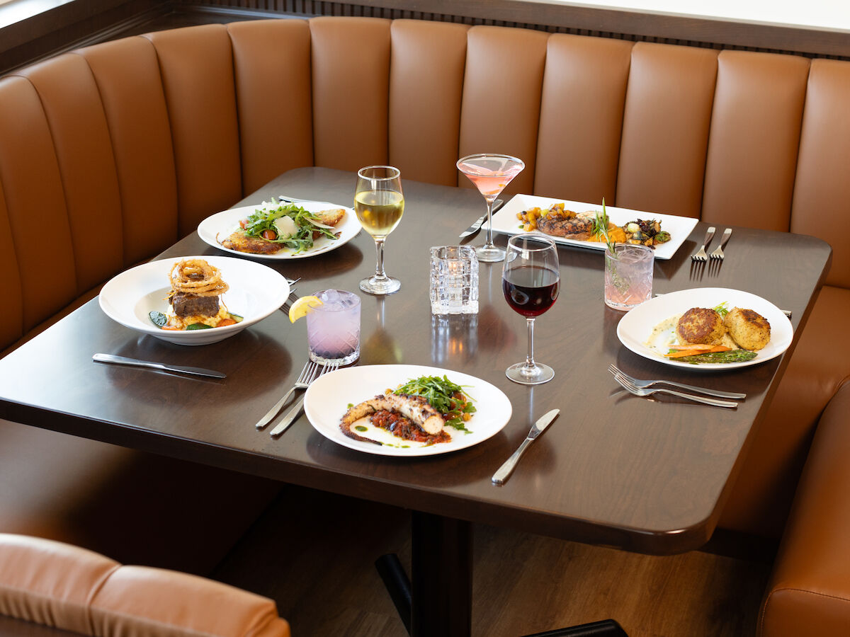 A restaurant booth with a wooden table set for four with various dishes, drinks, and utensils on top, in a cozy brown leather seating area.