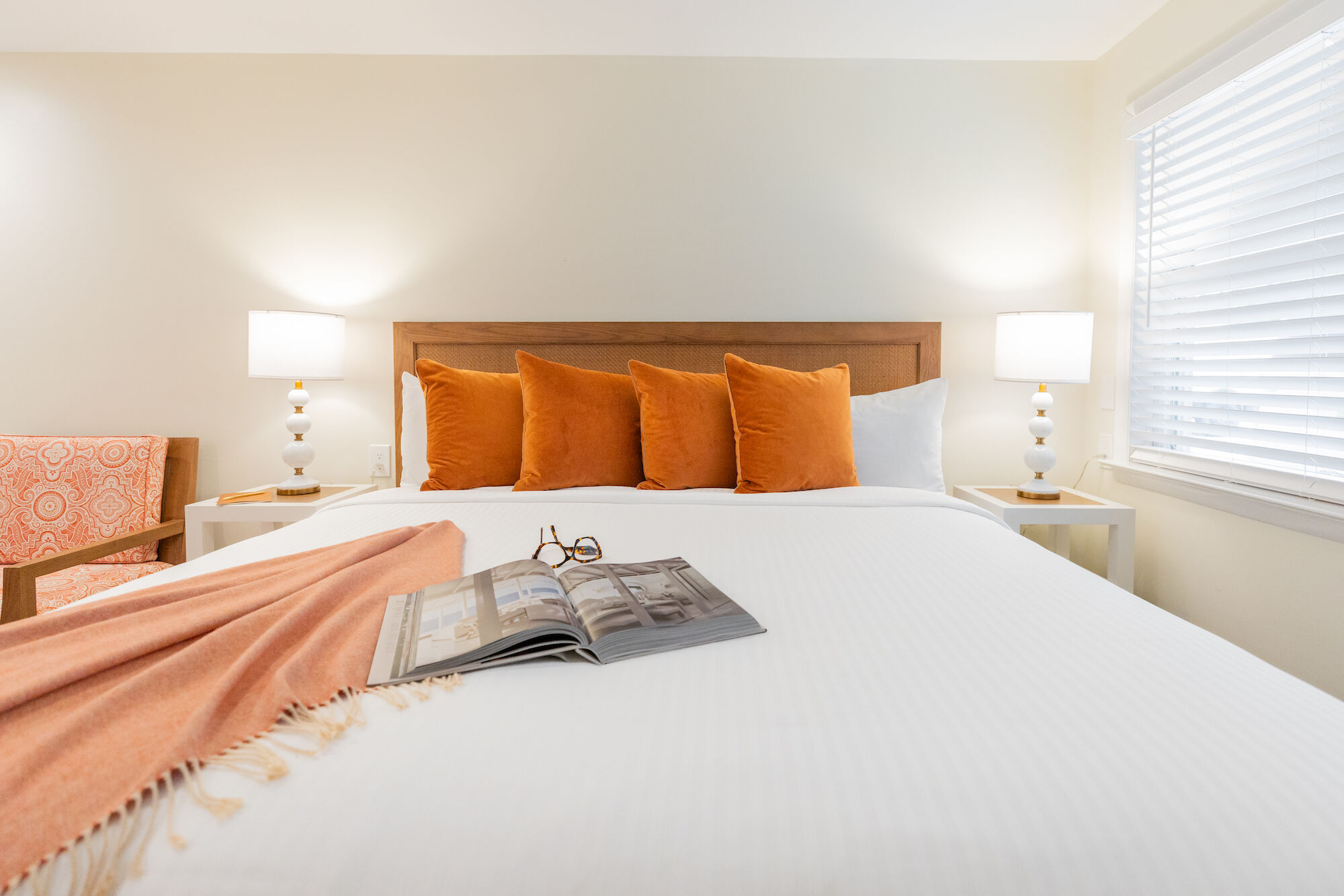 A neatly made bed with orange pillows, a peach blanket, an open book, and glasses on it. Two bedside lamps are lit in a cozy bedroom.