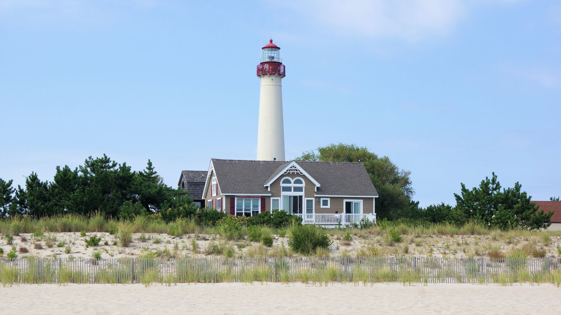 A lighthouse stands behind a charming house surrounded by greenery, with sand and a seagull in the foreground under a clear sky.