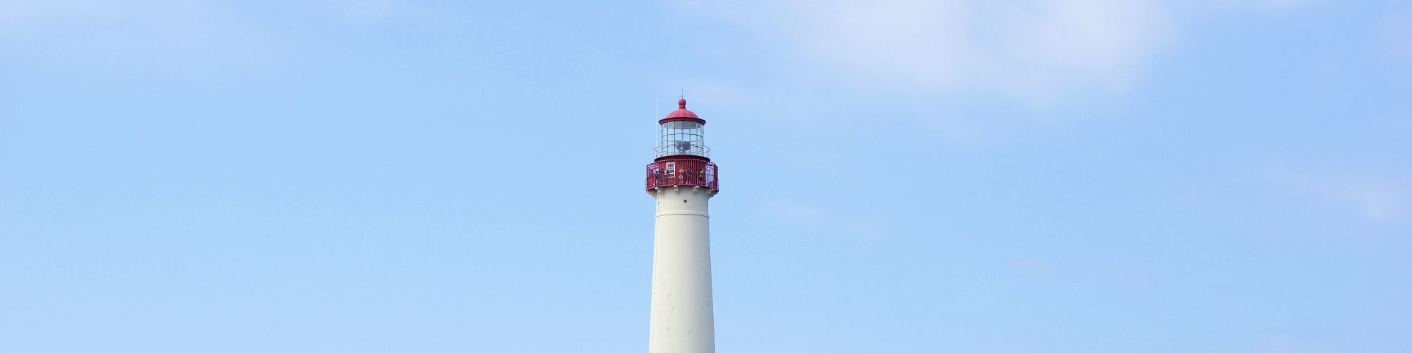 A lighthouse stands behind a charming house surrounded by greenery, with sand and a seagull in the foreground under a clear sky.