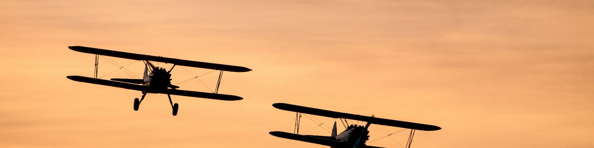 Two biplanes are silhouetted against a golden-orange sky, flying in close formation during sunset or sunrise, creating a picturesque scene.