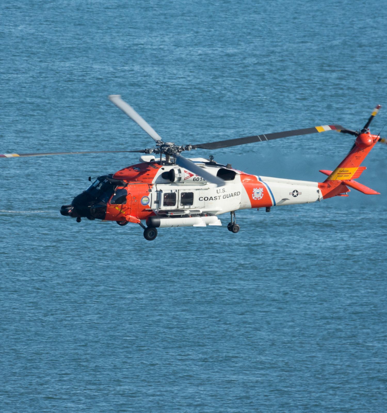 A U.S. Coast Guard helicopter flies over the ocean near a white sailboat, with both in the scene for a maritime context.