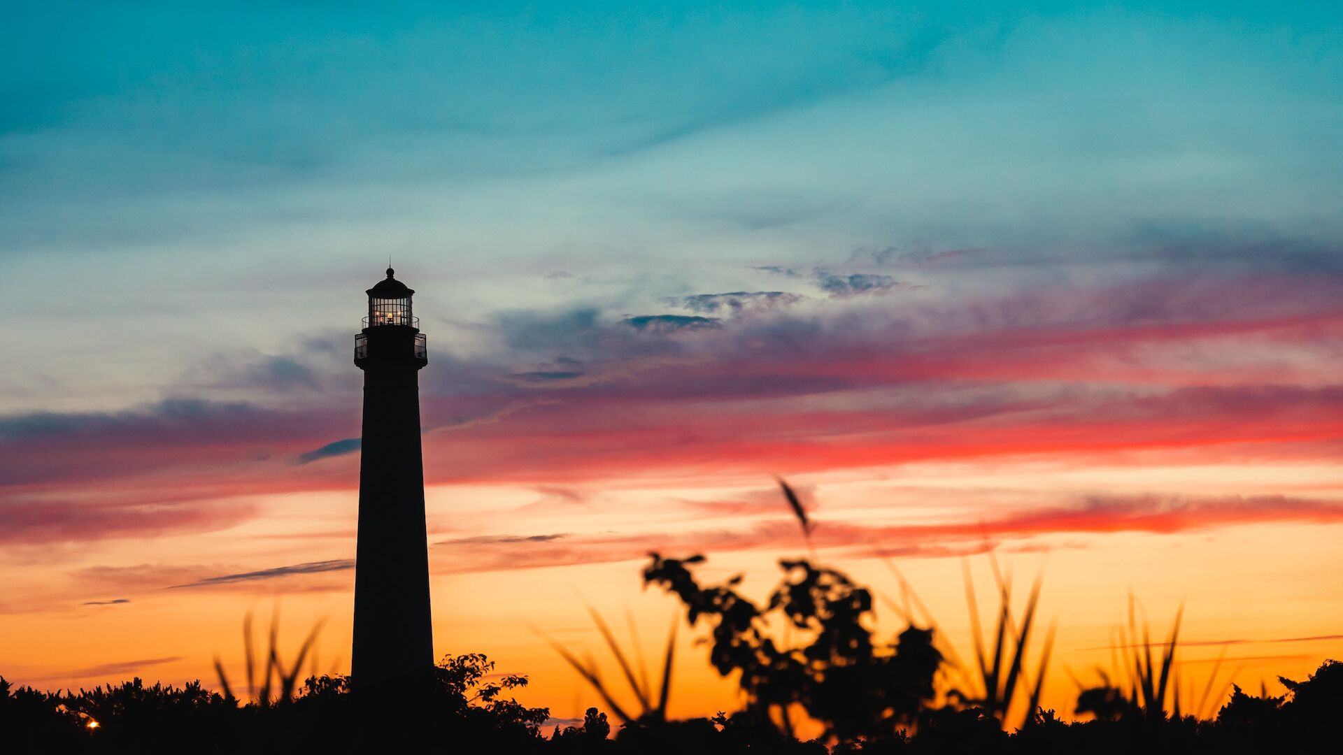 The image shows a lighthouse silhouetted against a vibrant sunset, with shades of orange, pink, and blue in the sky and foliage in the foreground.