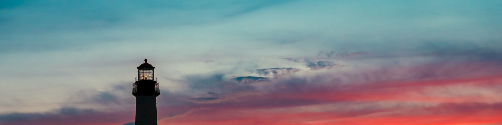 The image shows a lighthouse silhouetted against a vibrant sunset, with shades of orange, pink, and blue in the sky and foliage in the foreground.