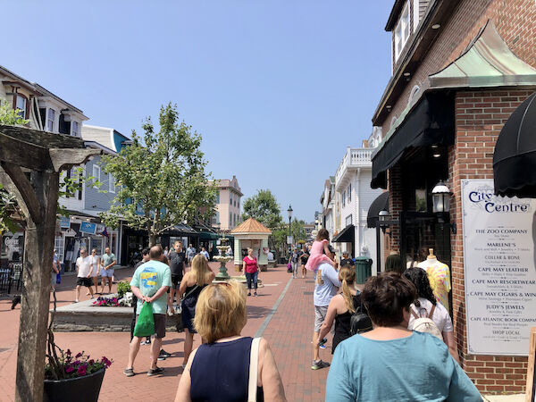 A bustling pedestrian street with people walking and shopping, surrounded by quaint shops and greenery, under a clear blue sky.
