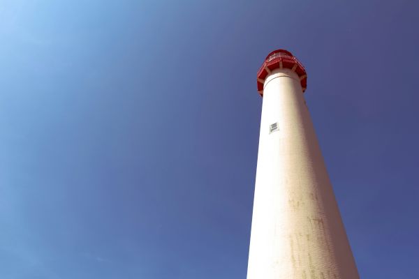 The image shows a tall lighthouse with a red top, viewed from below against a clear blue sky, appearing bright and majestic.