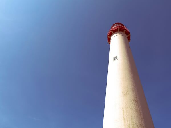 The image shows a tall lighthouse with a red top, viewed from below against a clear blue sky, appearing bright and majestic.