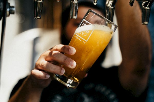 A person is holding a glass under a tap to pour a beer. The glass contains amber-colored beer with a frothy head forming at the top.