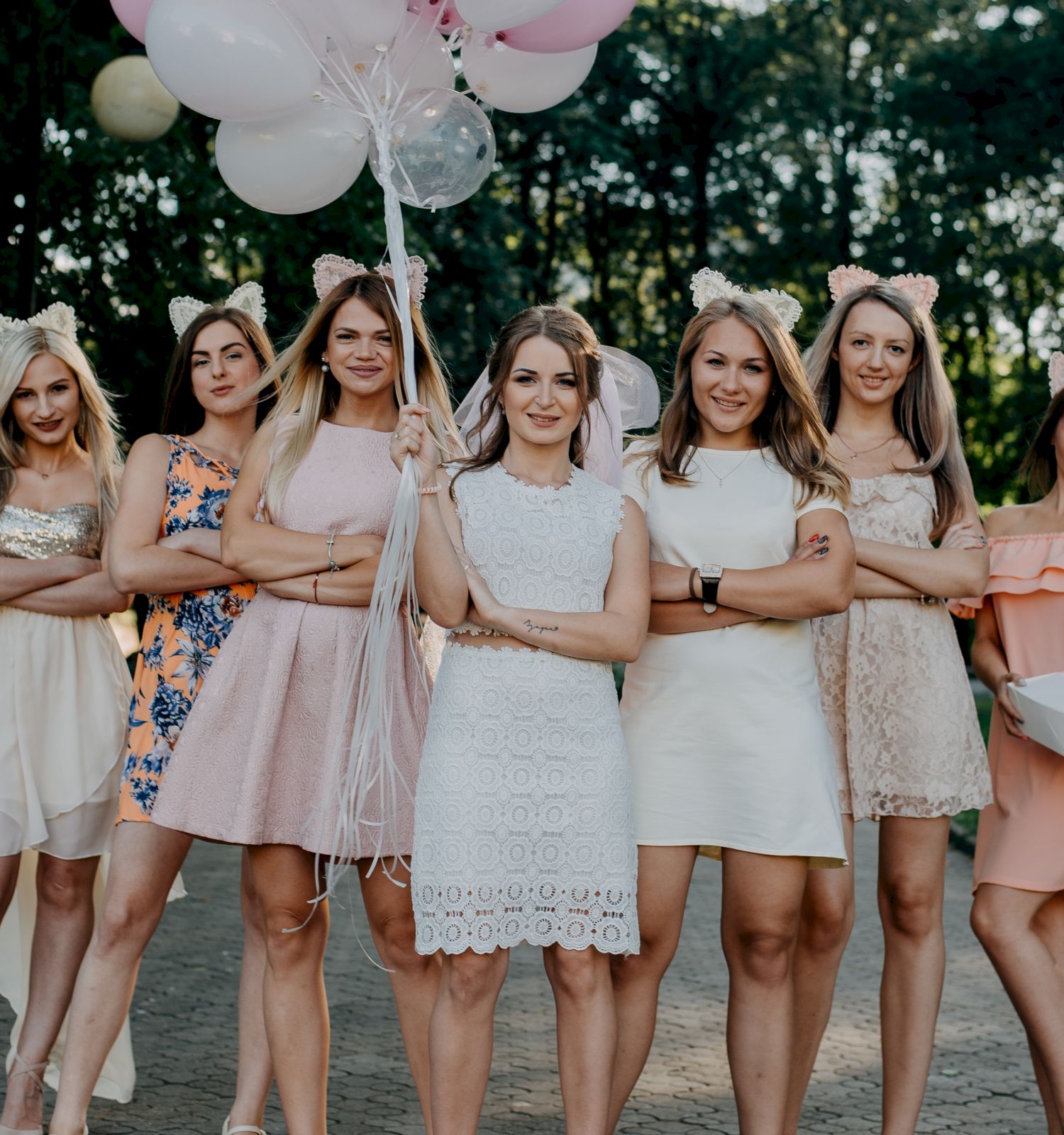 A group of women in dresses and bows pose outdoors; one holds balloons and another a gift box, standing on a tree-lined path.