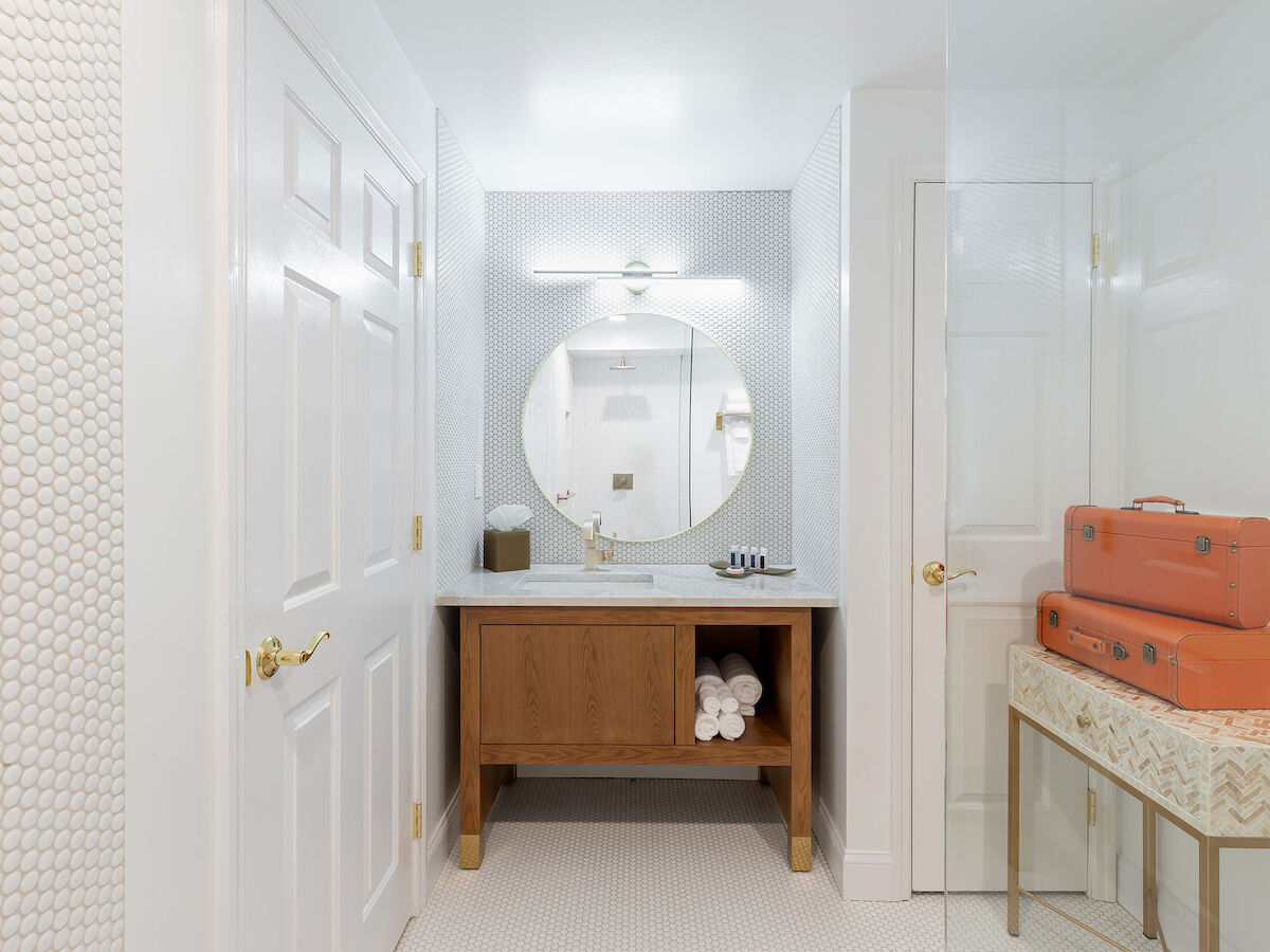 A bathroom with a round mirror, wooden vanity, rolled towels, and orange suitcases on a bench against a white, hexagon-tiled wall.