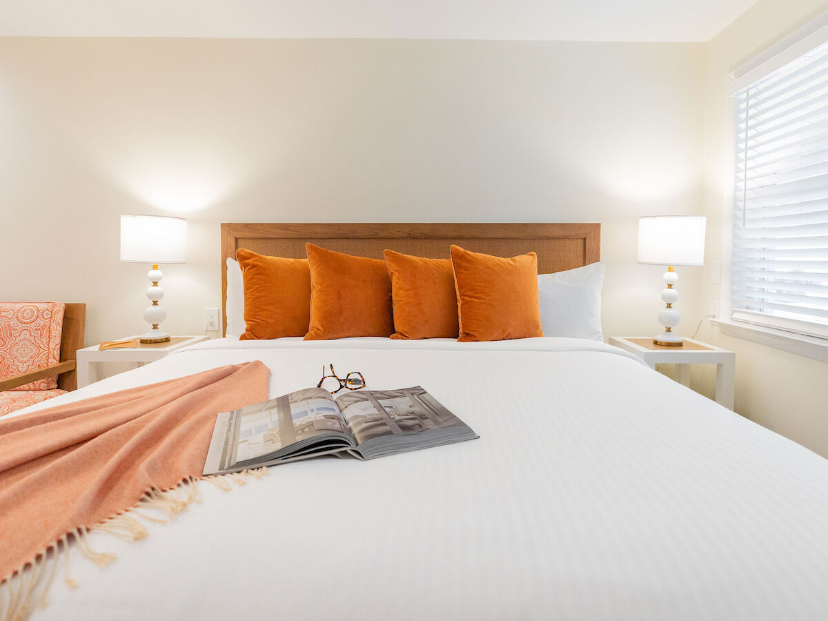 A neatly made bed with orange pillows, a blanket, and an open book on top; two side tables with lamps are situated beside it.