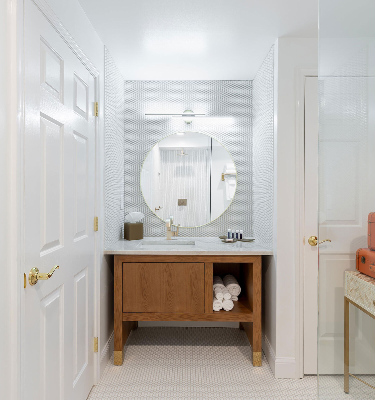 A bathroom with a round mirror, wooden vanity, towels, and two orange suitcases on a table, designed in a clean and minimalistic style.